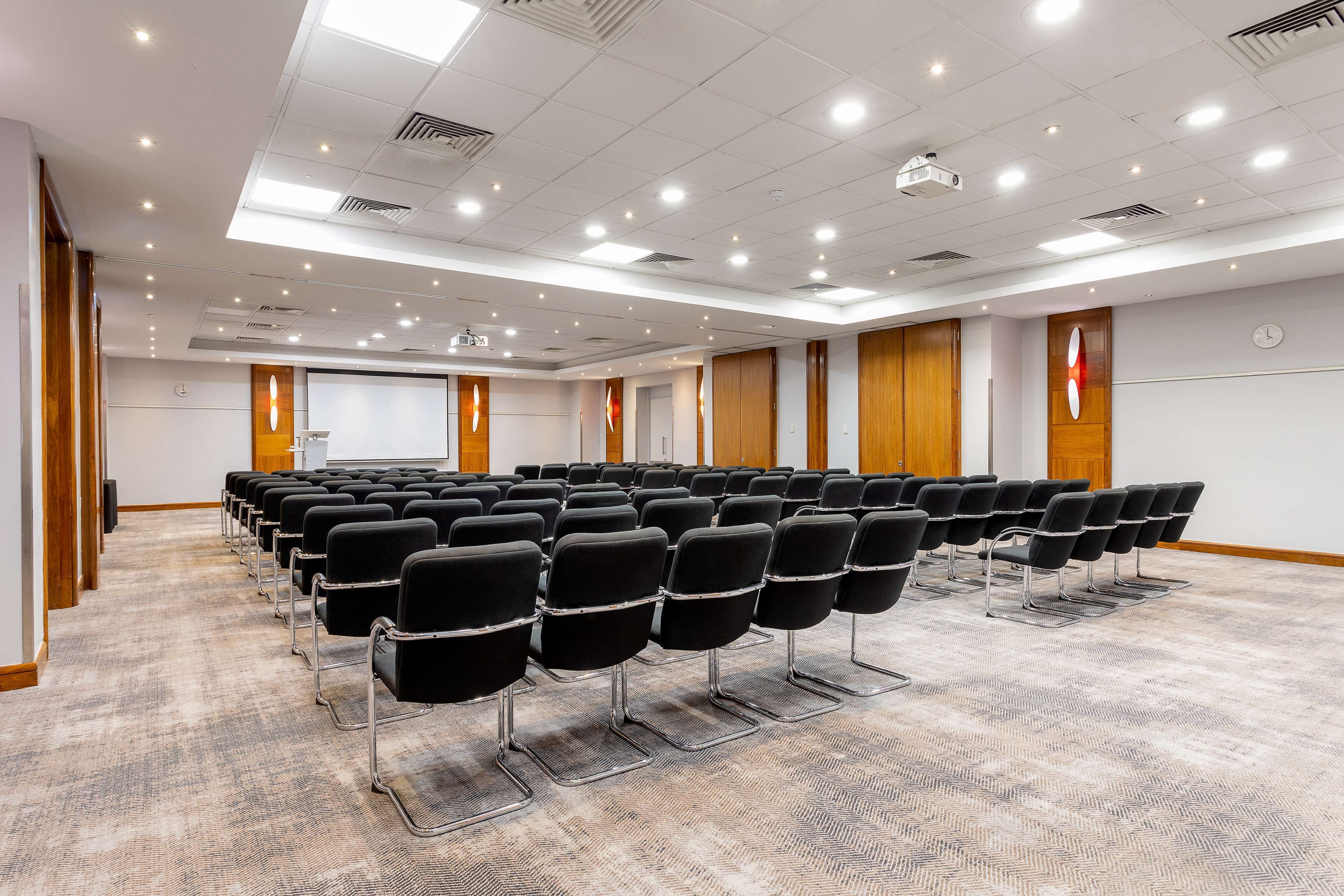 Une salle de conférence avec des rangées de chaises noires, un sol recouvert de moquette, des murs lambrissés et un écran de projection.