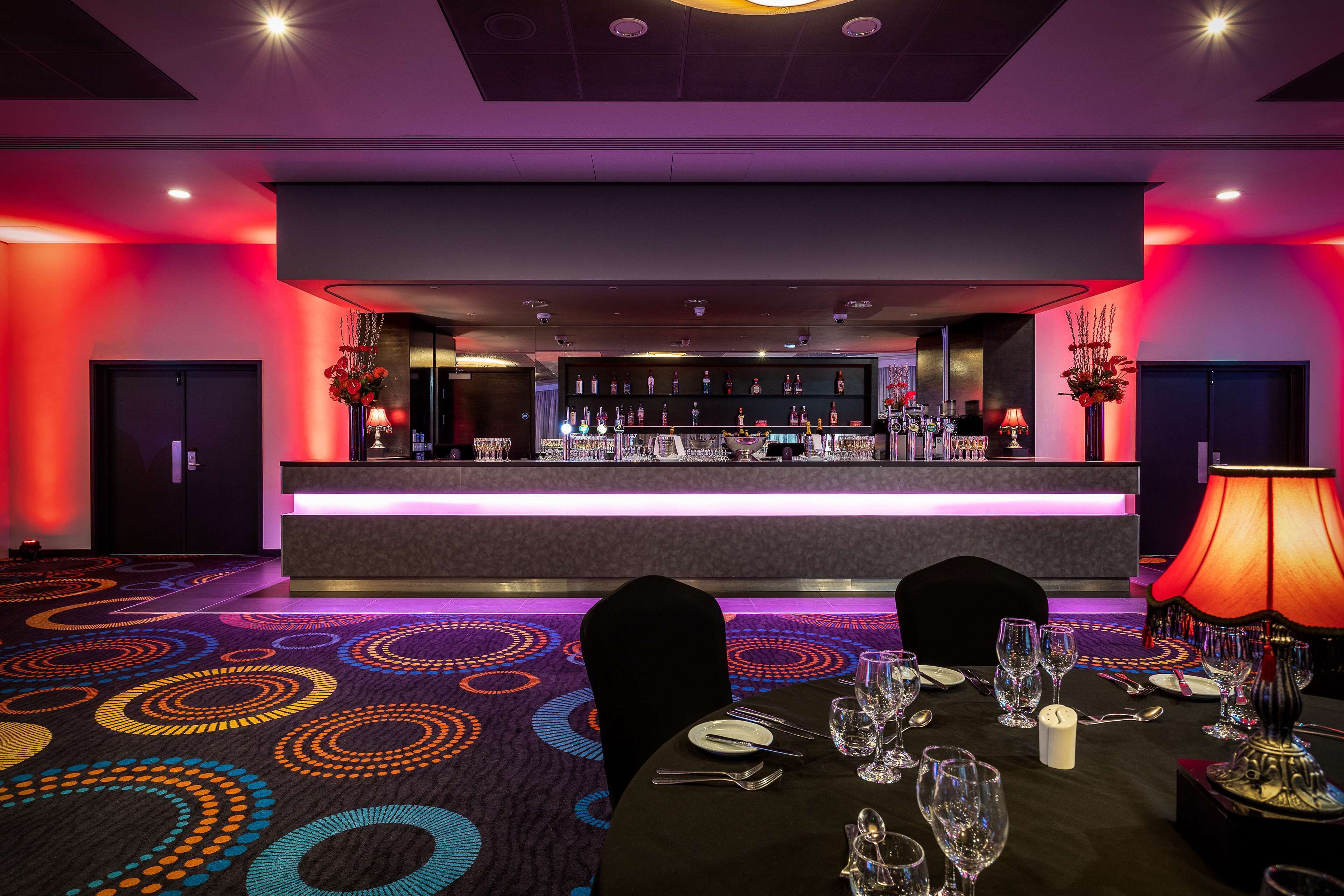 A restaurant with a bar, tables set with glasses and cutlery, and a patterned carpet.