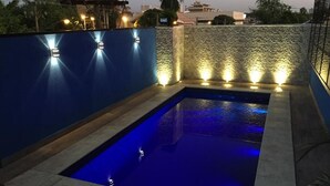 Una piscina con illuminazione blu, circondata da un muro in pietra e pannelli blu, con vista sullo skyline urbano.