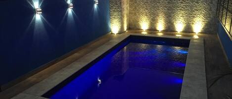 A swimming pool with blue lighting, surrounded by a stone wall and blue panels, with cityscape views in the background.