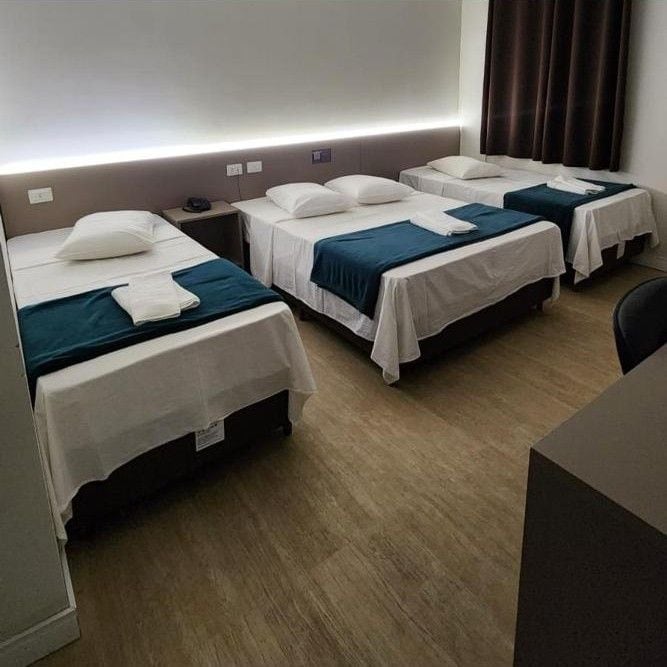 A hotel room with three single beds, each with a pillow and a blanket. There is a desk and a chair in the room.