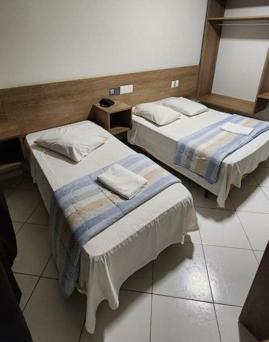 Une chambre d’hôtel avec deux lits simples, une tête de lit en bois, une table de chevet avec un téléphone et une petite étagère.