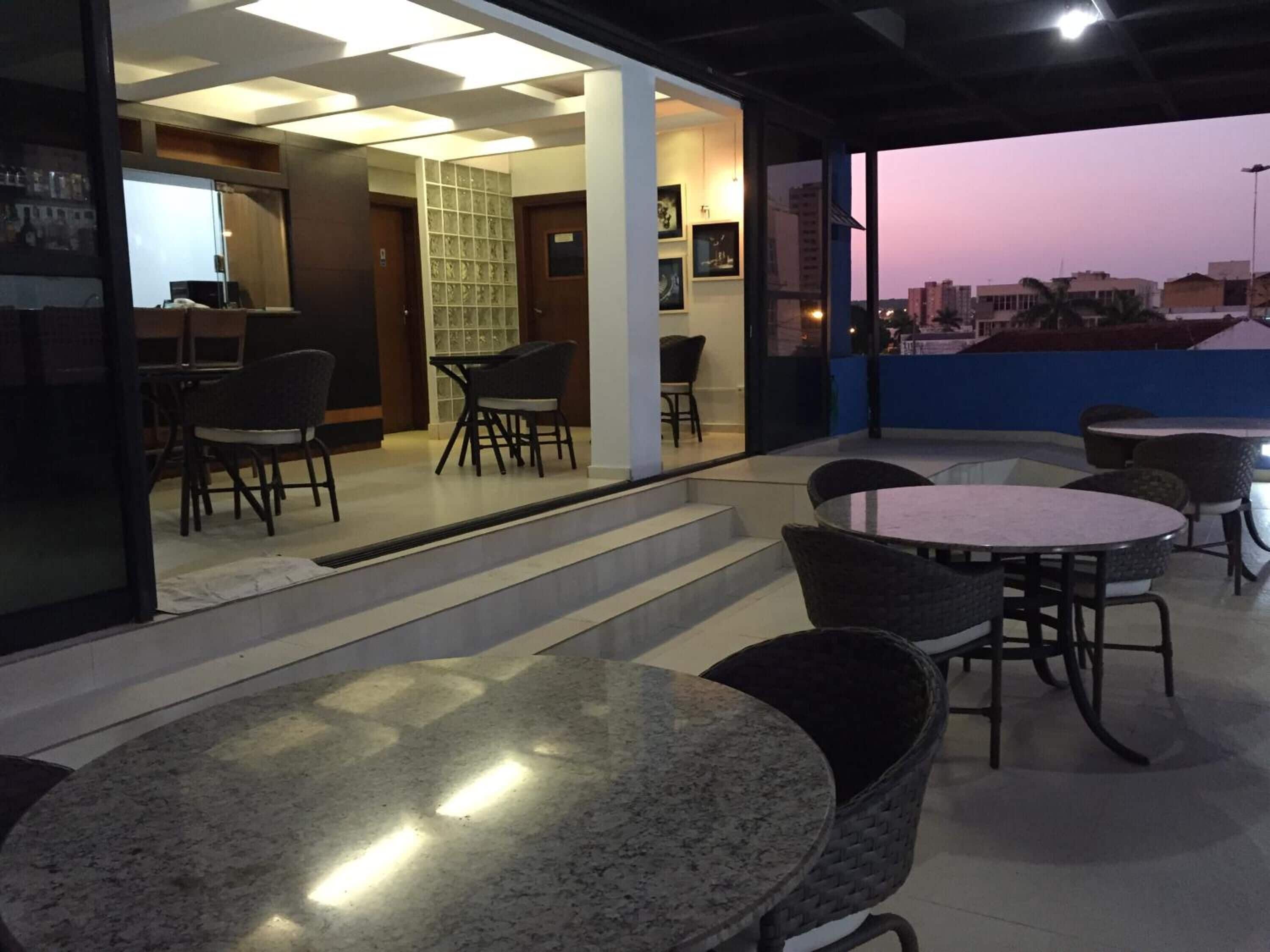 Un área de estar interior moderna con mesas y sillas, una escalera que conduce a un nivel superior y vistas de la ciudad al atardecer.