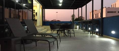 Un área de estar al aire libre con sillones reclinables y mesas, una sección cubierta con techo y vistas a edificios al fondo.