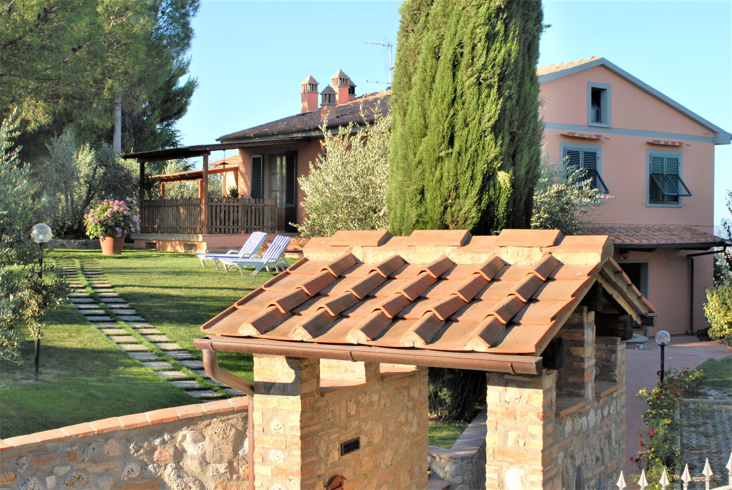 Une pergola en pierre surmontée d’un toit en tuiles, un mur en pierre et une pelouse bien entretenue agrémentée d’une terrasse en bois et de chaises longues.
