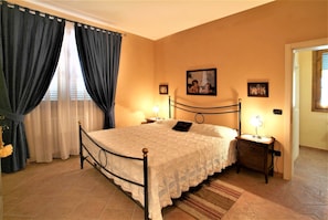 A bedroom with a bed, curtains, a nightstand with a lamp, a picture on the wall, and a door.