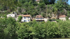A hillside with stone buildings, some with blue shutters, surrounded by dense green foliage and a body of water in the foreground.
