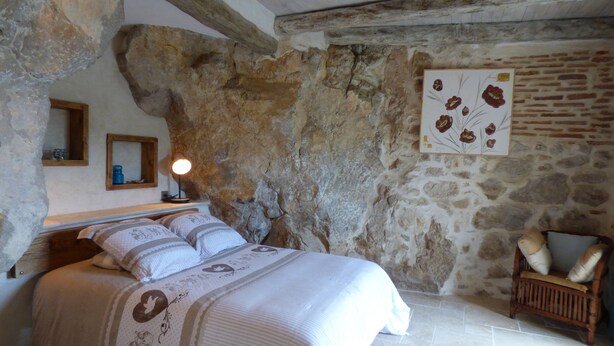 A bedroom with a stone wall, a bed with patterned bedding, a wooden nightstand, a wall-mounted lamp, and a chair.