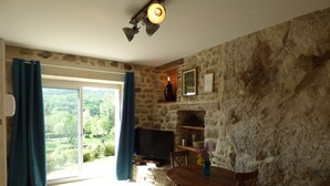 A room with a stone wall, a wooden dining table with chairs, a TV, and a view of greenery outside.