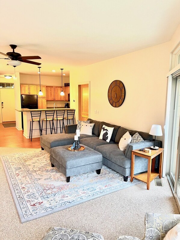 A modern living room with a sectional sofa, ottoman, and a small side table. There is a dining area with a bar and chairs in the background.