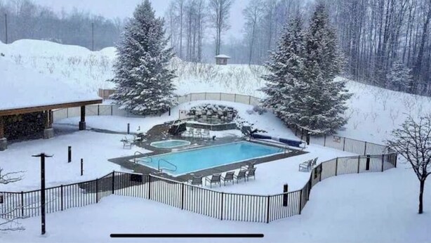Une piscine extérieure entourée de neige, avec une terrasse délimitée par une clôture, des chaises longues et un bâtiment en arrière-plan.