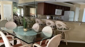 A dining area with a glass table surrounded by wicker chairs with orange cushions, a kitchen with wooden cabinets and white appliances, and a large mirror reflecting the space. - Desired Resort on Hilton Head Island (3 BD, 3BA Condo near beach) (Hilton Head)
