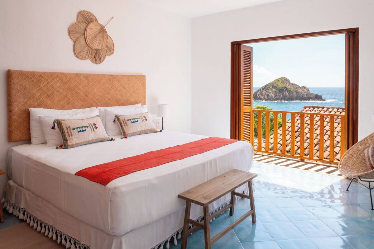 Une chambre avec un grand lit, une tête de lit en bois, un plaid rouge et une vue sur l’océan par une porte ouverte.