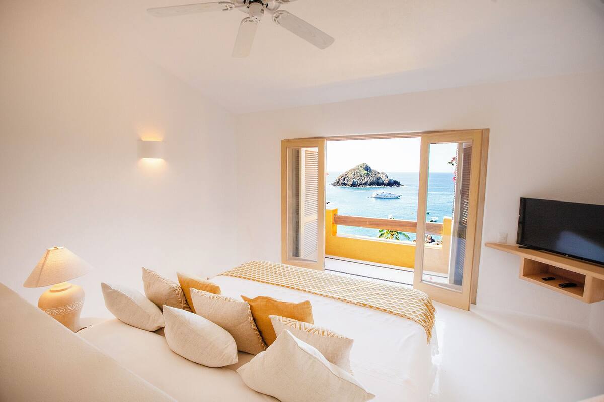 Une chambre d’hôtel avec un lit, des oreillers, une télévision, un ventilateur de plafond et une vue sur la mer.