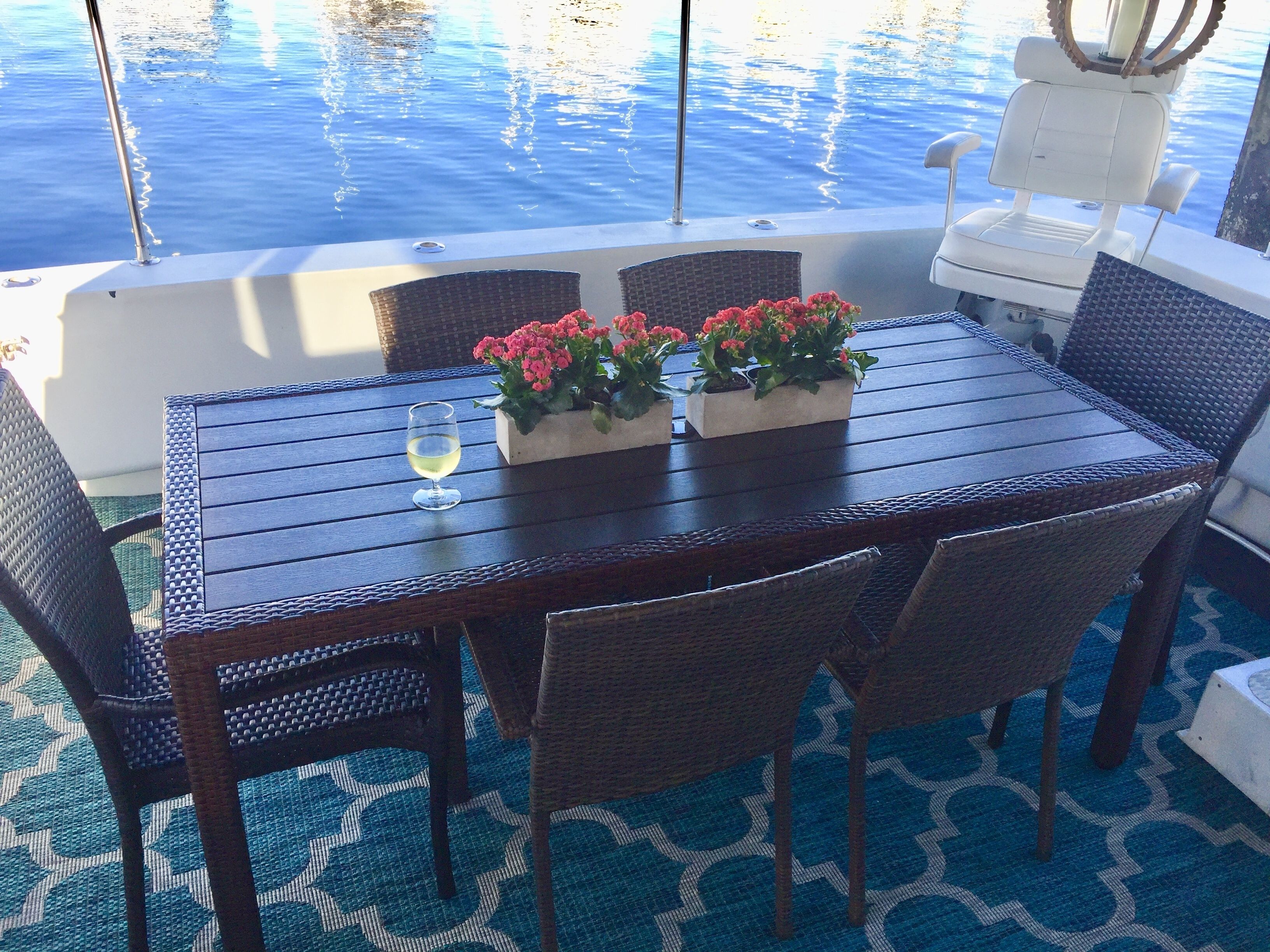 A dining table with wicker chairs, two flower pots, and a glass of white wine on a boat deck with a view of the water.