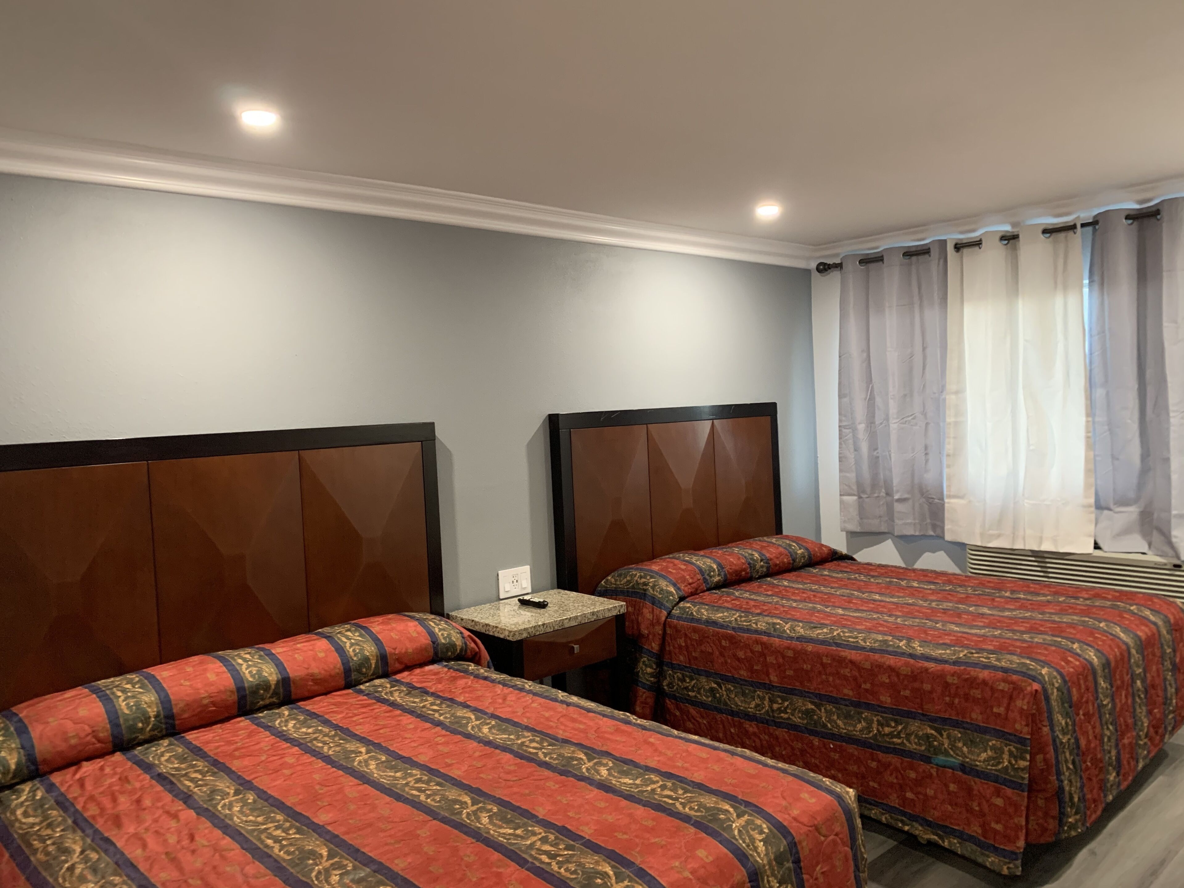 A hotel room with two beds, a small table, a power outlet, and a window with curtains.