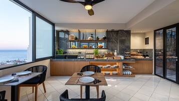 A modern kitchen with a view of the sea, featuring a dining area with a table and chairs, and a breakfast bar with various items.