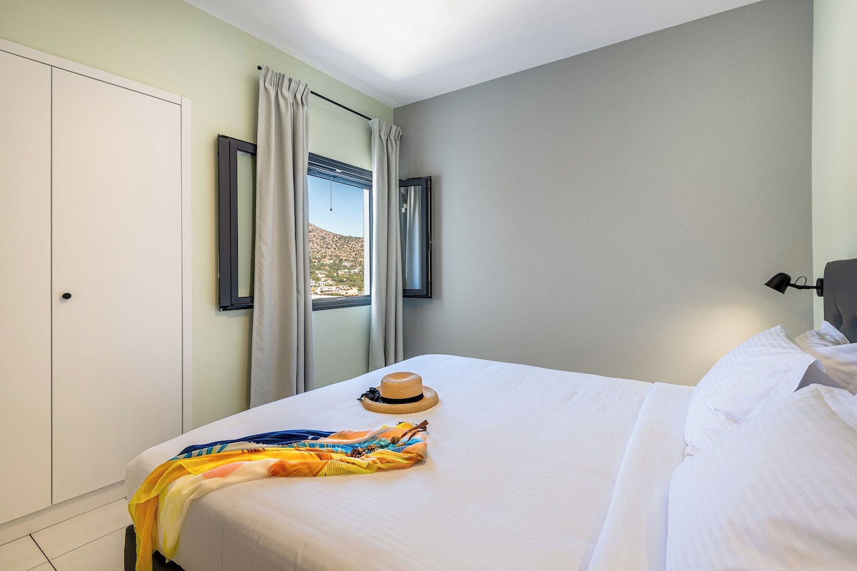 A neatly made bed with a hat and a colorful blanket, a window with curtains, and a view of a mountainous landscape.