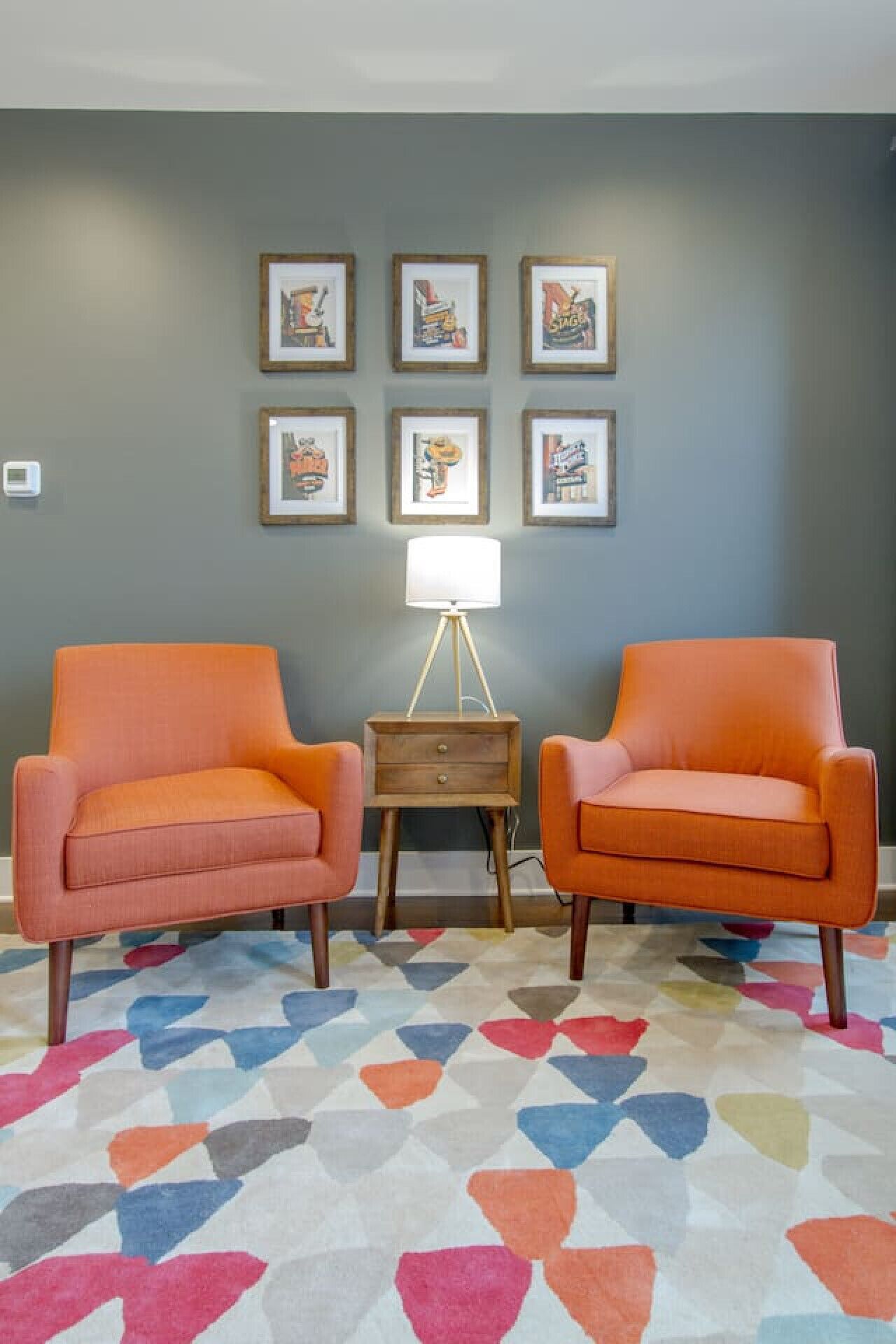 A room with two orange armchairs, a small wooden nightstand with a lamp, and six framed posters on the wall.