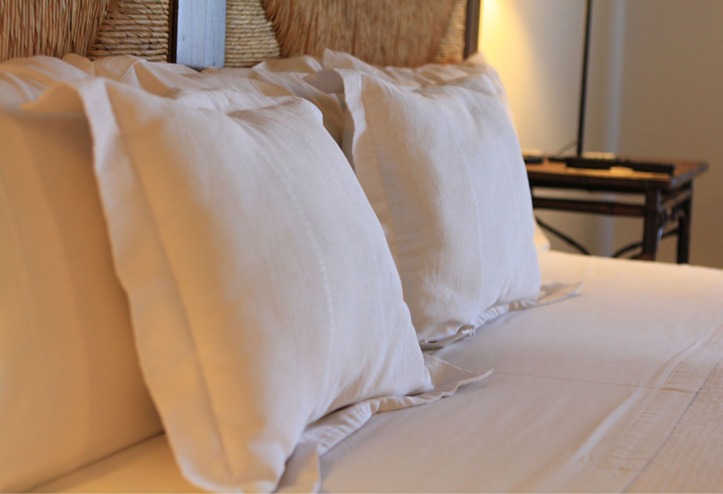 Una cama con sábanas y almohadas blancas, un cabecero tejido y una mesita de noche con una lámpara.