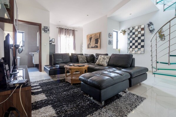 A modern living room with a sectional sofa, a coffee table, and a TV. There is a staircase with glass railings and a decorative wall piece.