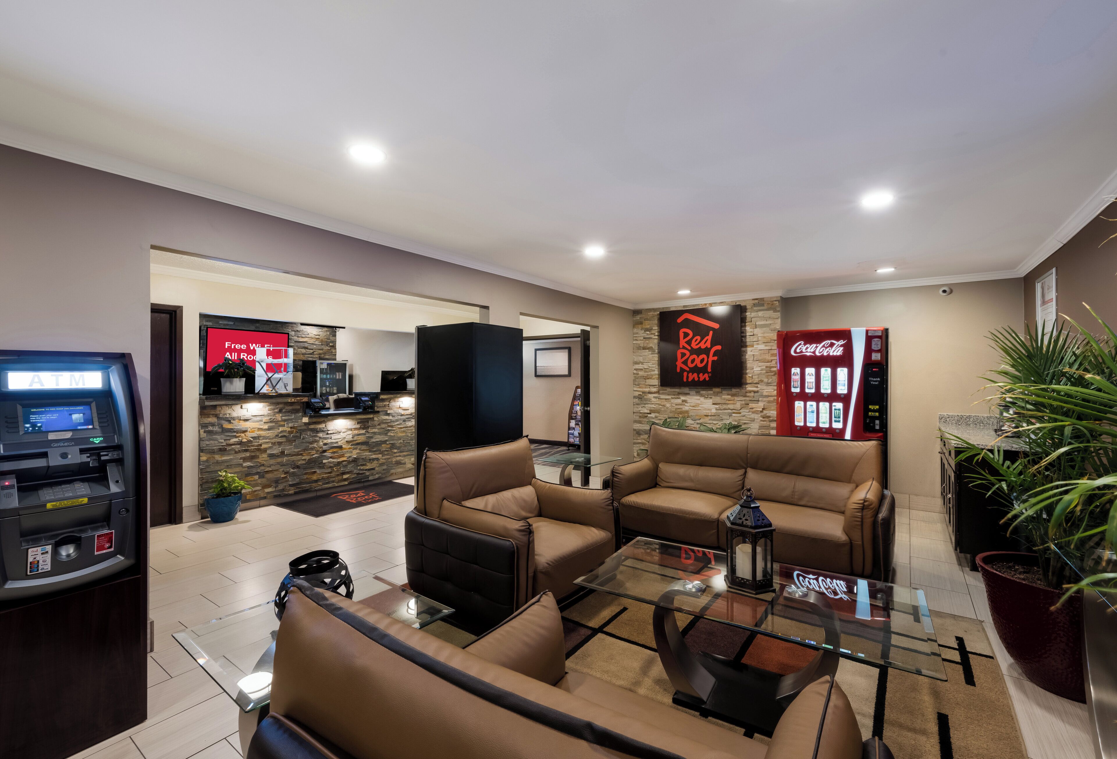 A lobby with leather sofas, a glass coffee table, an ATM, a Red Roof Inn sign, a Coca-Cola vending machine, and a stone wall.
