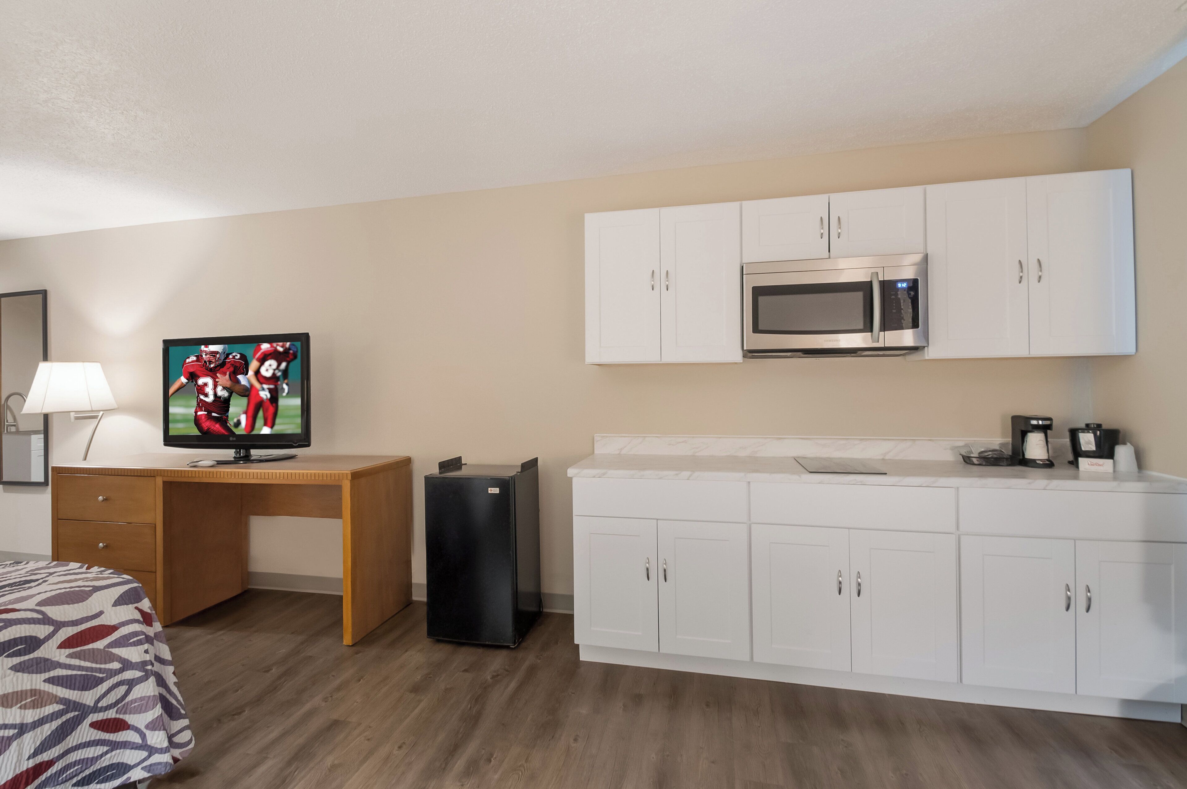 Habitación de hotel con cama, escritorio con televisor, refrigerador, microondas y kitchenette con alacenas.