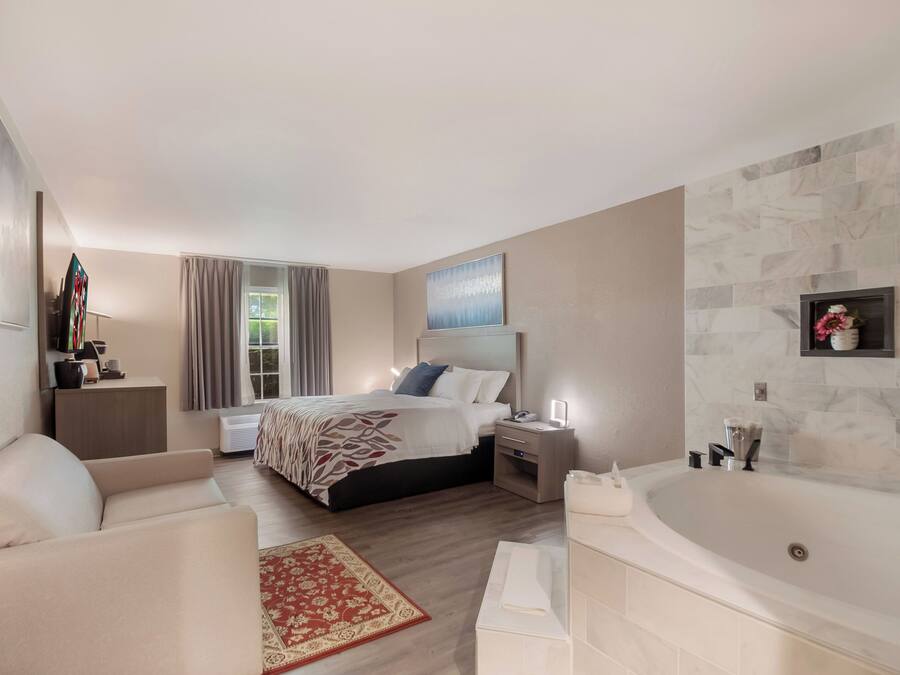 Una habitación de hotel moderna con cama, zona de estar, baño con bañera y un ventanal con cortinas.