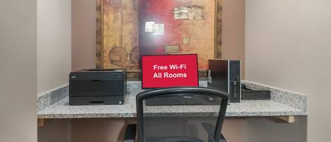 A workspace with a desk, two printers, a chair, and a wall-mounted sign indicating free Wi-Fi availability.