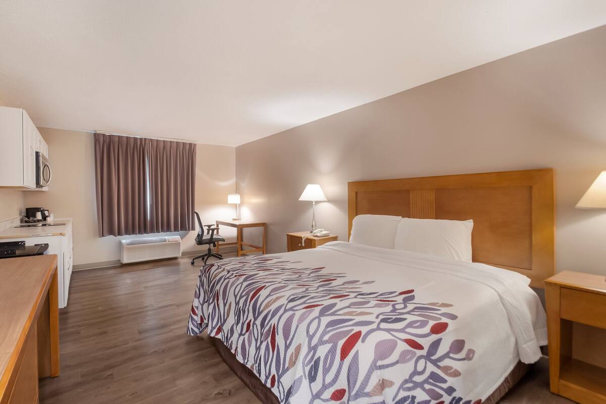 A hotel room with a bed, a desk, a chair, a lamp, a nightstand, and a window with curtains.