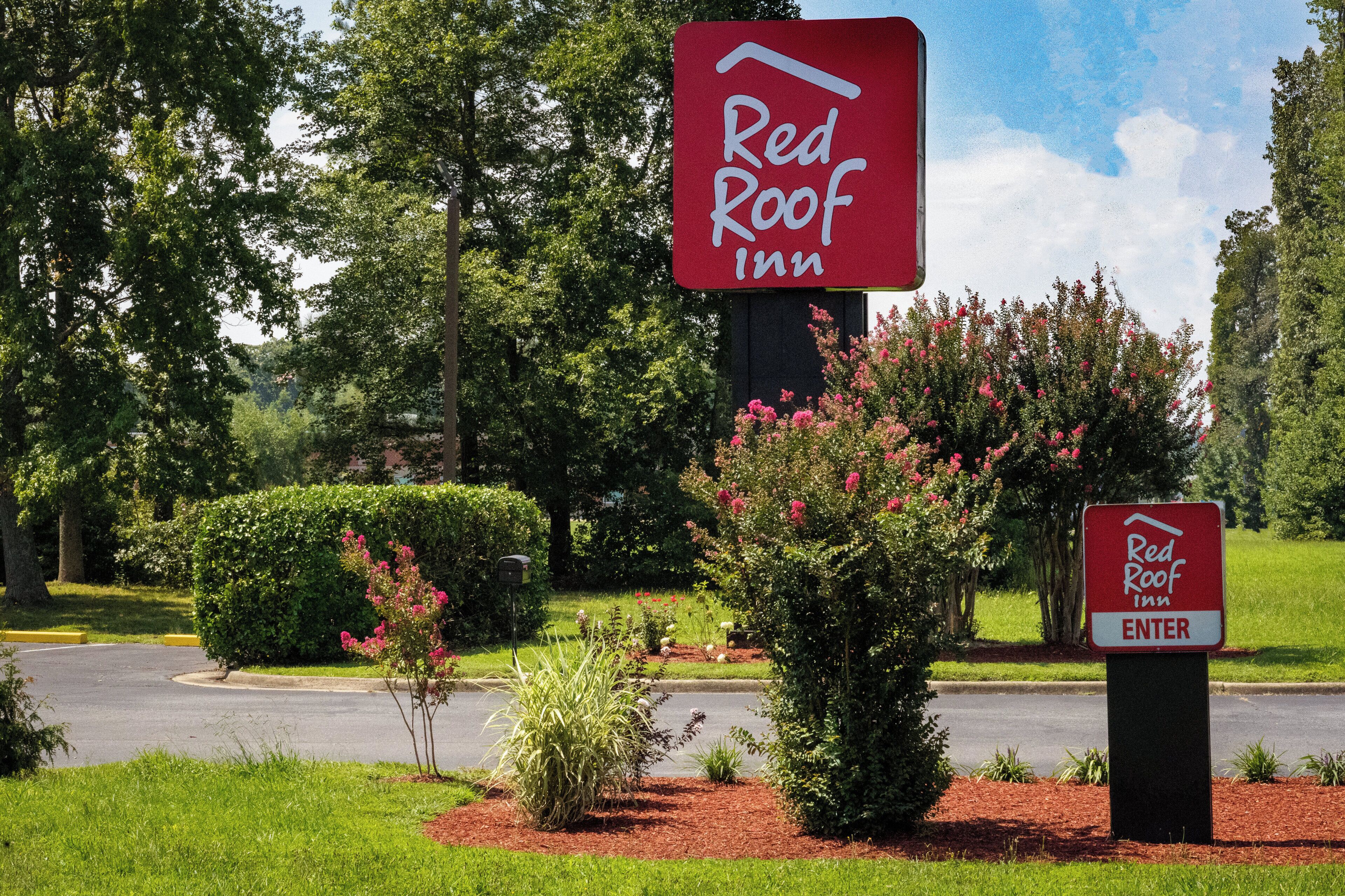 Señal de Red Roof Inn con un cartel que dice 'INGRESA', rodeada de follaje y una carretera.