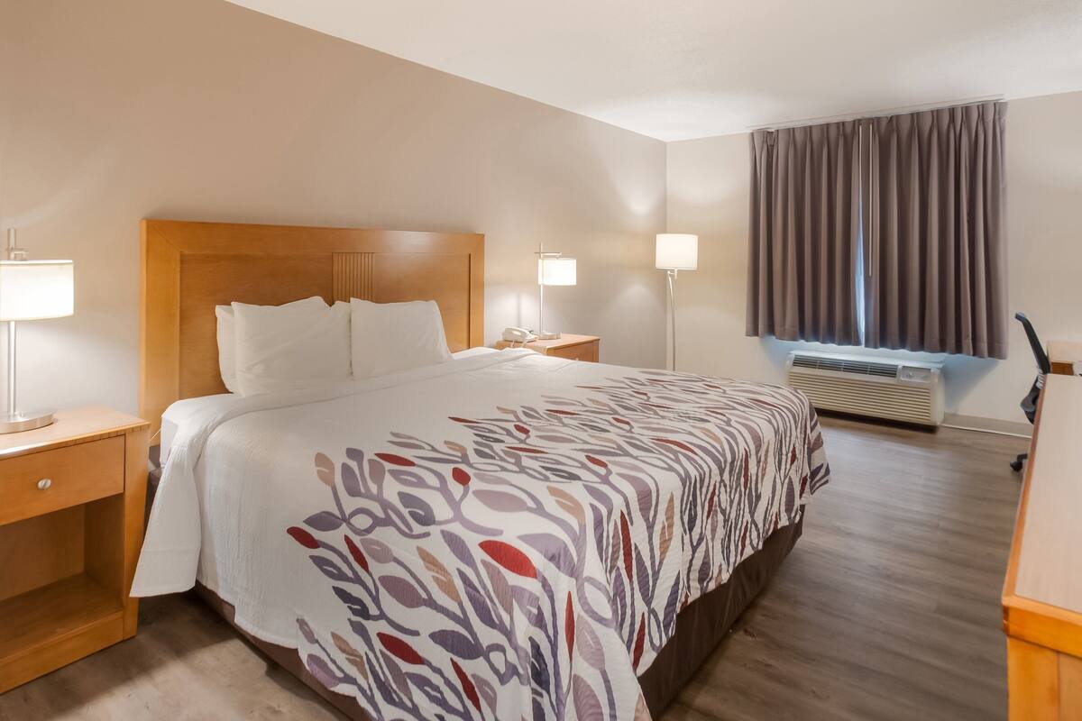 A hotel room with a large bed, bedside tables with lamps, a window with curtains, and an air conditioning unit.