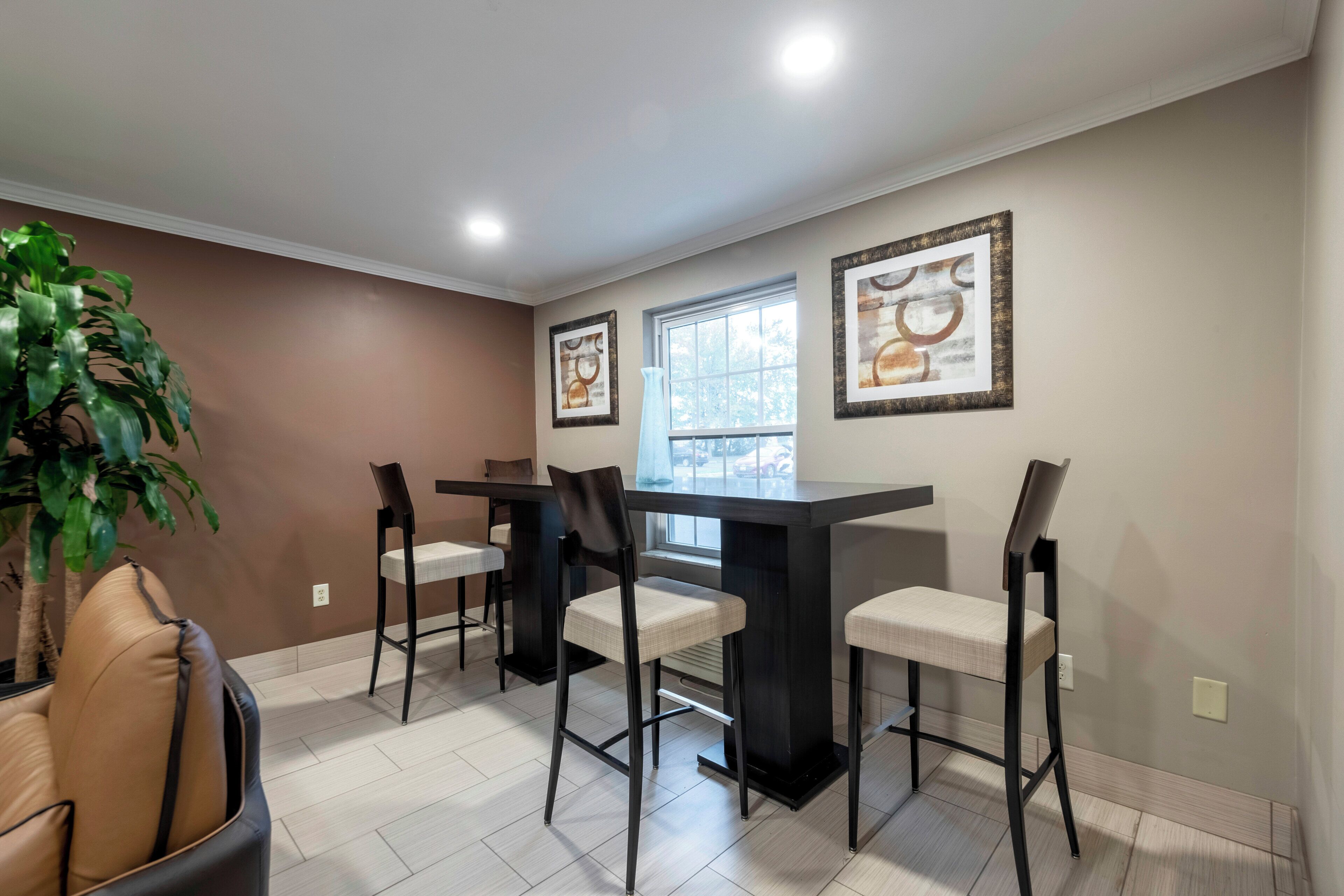 A dining area with a high table and four chairs, a potted plant, and two framed artworks on the walls.