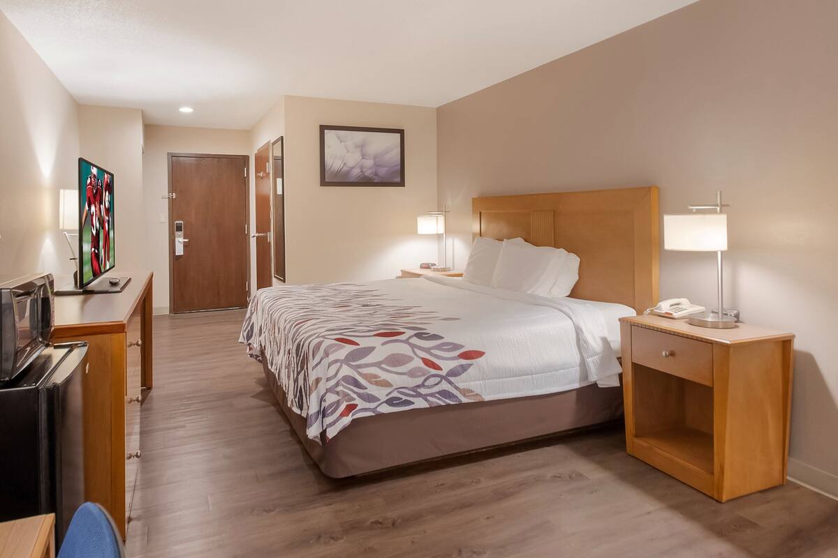 A hotel room with a bed, bedside lamps, a desk with a TV, a refrigerator, and a door.