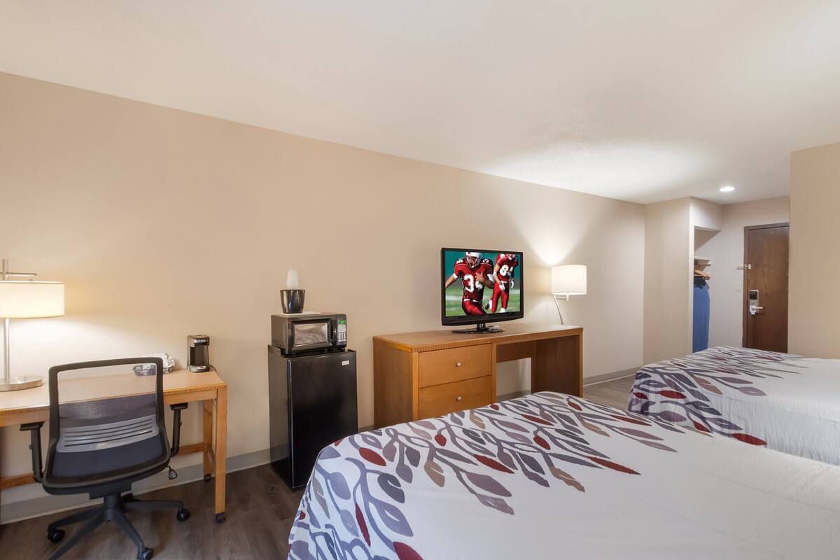A hotel room with a desk, a television, a refrigerator, a microwave, a chair, and two beds.