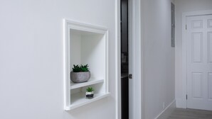 Un couloir aux murs blancs, avec une porte blanche et une étagère sur laquelle sont posées deux plantes en pot.