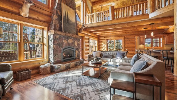 A log cabin interior with a stone fireplace, a large sofa, and a dining area. - Fireside Retreat: Highlands Log Home, Hot Tub & Fireplace, Peak Views! (Breckenridge)