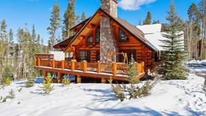 A log cabin with a stone chimney, wooden deck, and snow-covered surroundings. - Fireside Retreat: Highlands Log Home, Hot Tub & Fireplace, Peak Views! (Breckenridge)