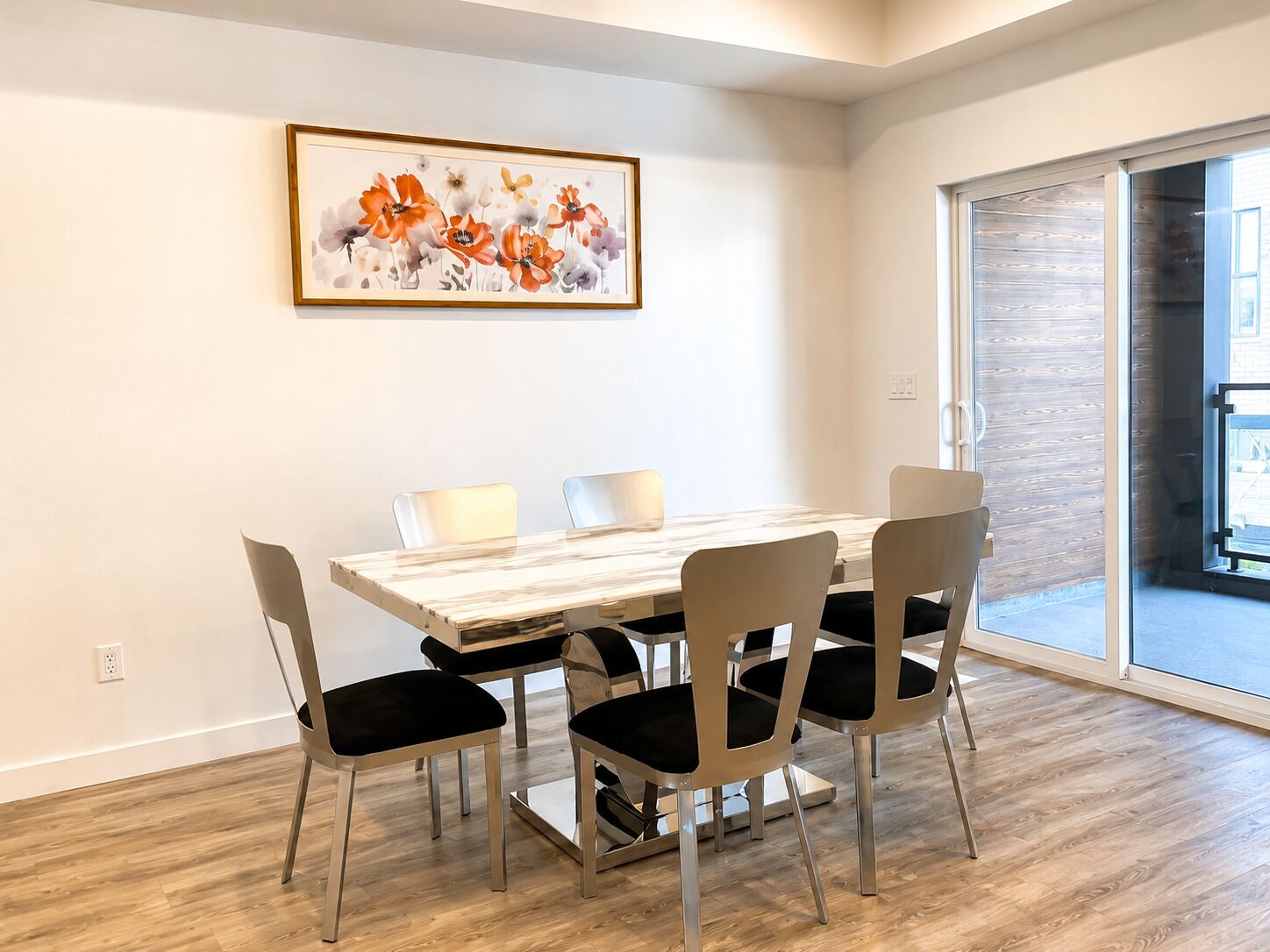 Een eethoek met een houten tafel en vier stoelen, een wanddecoratie met bloemen in een lijst, en een schuifdeur naar een terras.