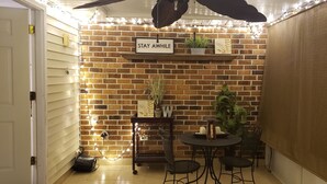 Ein gemütlicher Winkel mit einer Ziegelwand, einem Schild mit der Aufschrift 'STAY AWHILE', einem kleinen Tisch mit Stühlen und Lichterketten.