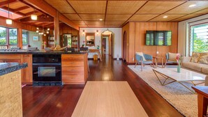 Una cocina amplia con armarios de madera, horno empotrado y una isla grande. Hay un comedor con sofá y sillas, y un televisor montado en la pared.