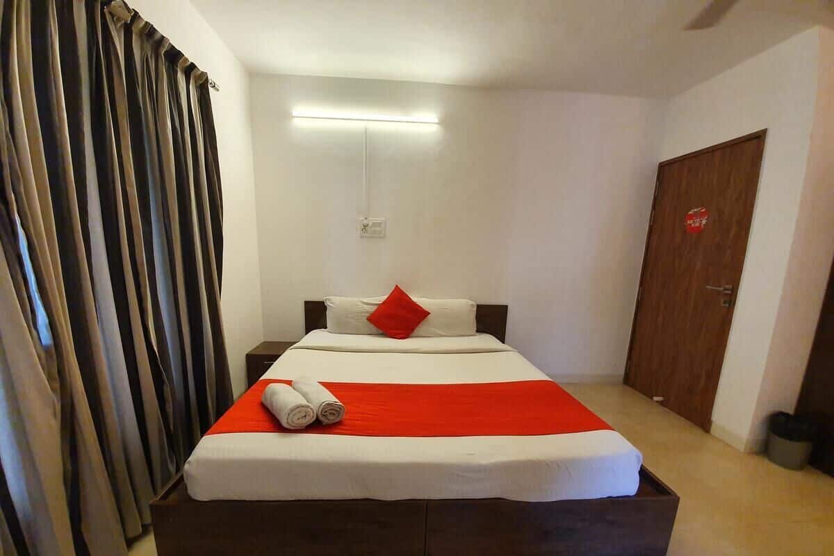 A neatly made bed with a red blanket, white pillows, and a red accent pillow. There is a wooden headboard, a bedside table, and a wall-mounted light fixture.