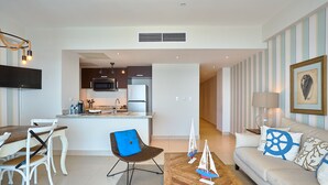 Una sala de estar moderna con un sofá, una mesa de centro y sillas. Se ve un área de cocina con un microondas y un refrigerador al fondo.