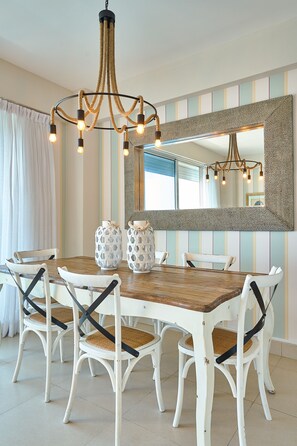 A dining area with a wooden table and white chairs with black frames. There is a chandelier above the table and a large mirror on the wall. - Marvelous Ocean Views, Harmony 6FLR Apt at Las Olas Metro Country Club - Unit #2 (Playa Juan Dolio)