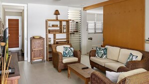 Un salon avec des meubles en osier, un buffet en bois et une grande fenêtre équipée de stores.