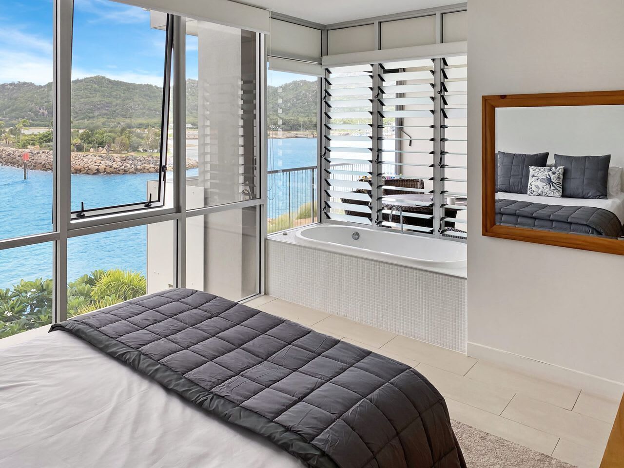 A bedroom with a large bed, a view of a body of water, a bathtub, and a window with blinds.