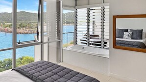 Une chambre avec un grand lit, une vue sur un plan d’eau, une baignoire et une fenêtre avec des stores.