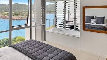 A bedroom with a large bed, a view of a body of water, a bathtub, and a window with blinds.