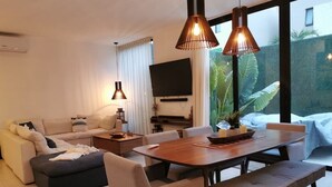 Un salon moderne avec un canapé, une table basse et une table à manger avec des bancs. Un téléviseur à écran plat est fixé au mur, et une grande fenêtre offre une vue sur la verdure environnante.