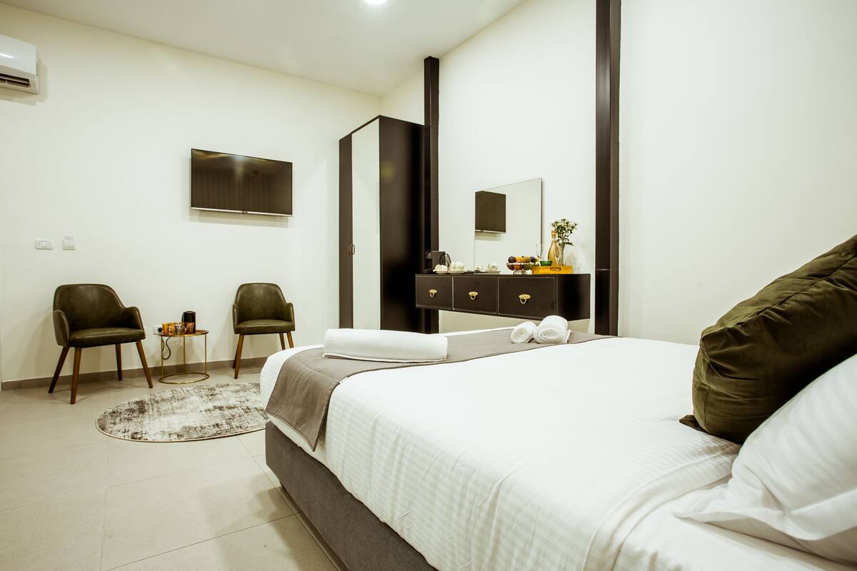 A modern hotel room with a large bed, two armchairs, a small side table, a flat-screen TV, a dresser with a mirror, and a large window.